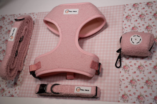 A set of pink and blue pet harness and leash accessories displayed on a floral patterned background, including a harness, leash, collar, and poop bag.